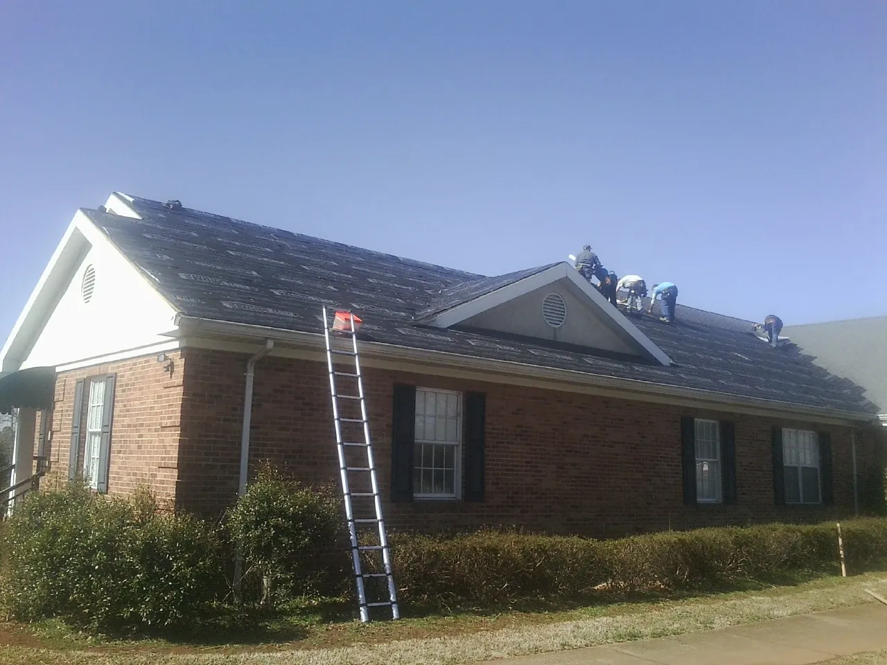 Crew tearing shingles off a brick ranch roof with a ladder set — Metal Roof Repair work by our crew in Gilmanton