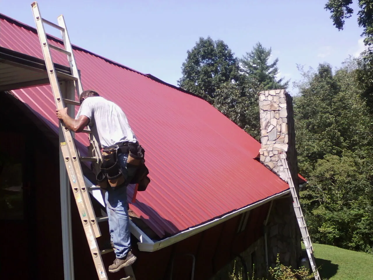 Roofer on a ladder beside a red metal roof and stone chimney — Soffit & Fascia Repair by Roofing Brothers in Gilmanton